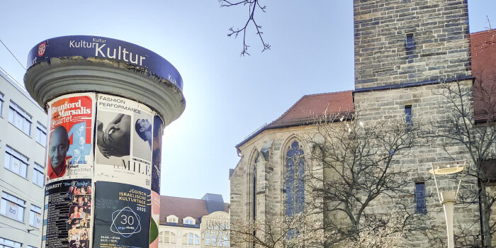 eine mit Plakaten beklebte Litfaßsäule vor einer Kirche