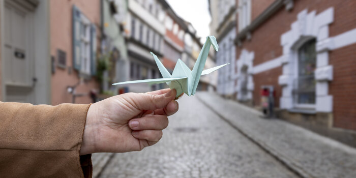 eine Hand hält einen blauen Papierkranich, im Hintergrund die Erfurter Krämerbrücke