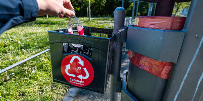 Ein Kasten ist an einem Mülleimer angebracht, eine Hand stellt eine Flasche hinen