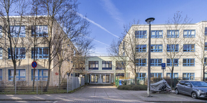 Ein Schulgebäude. Zwei dreistöckige Gebäude werden durch einen einstöckigen Mittelbau verbunden. 