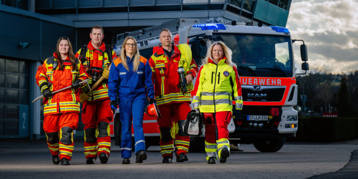 Fotomontage: Drei Frauen und zwei Männer in verschiedenen Uniformen laufen vor einem Feuerwehrfahrzeug. 