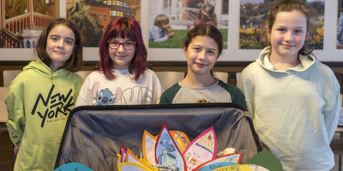 Vier Kinder stehen hinter einem offenen Koffer. Darin sind gemalte Bilder und Plakate.