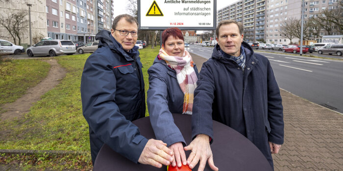 Zwei Männer und eine Frau stehen vor einer digitalen Anzeigetafel und halten ihre Hände über einen Buzzer. 