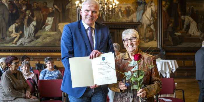 Ein mittelalter Mann hält einen Ehrenbrief für eine ältere Frau, die wiederum eine Blume in der Hand hat. 