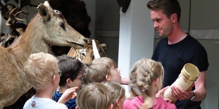 ein Mann hält eine Art Stoßzahn in der Hand und zeigt ihn mehreren Kindern