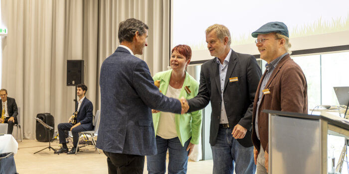 vier Personen, zwei davon reichen sich die Hand zur Gratulation