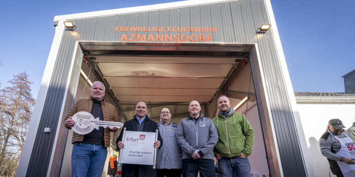 Fünf Männer stehen vor einer Fahrzeughalle. Einer hält ein Schild und einer einen überdimensionalen Schlüssel.