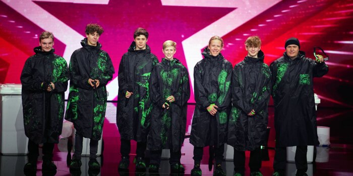 Sieben Personen in schwarz-grünen Umhängen stehen auf einer Fernsehbühne