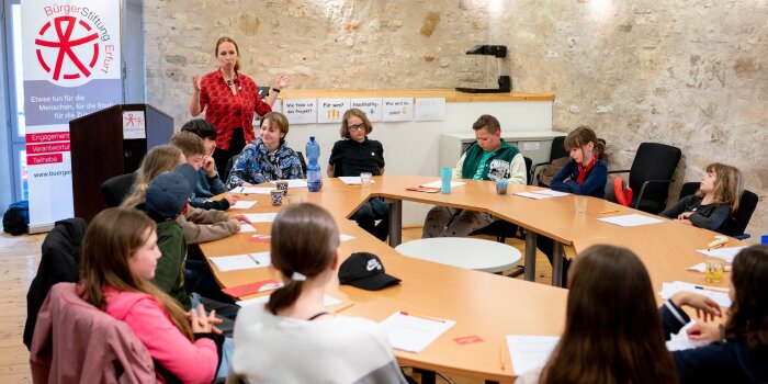 Mehrere Kinder sitzen an einem runden Tisch. Im Hintergrund steht eine Frau an einem Pult und spricht. 