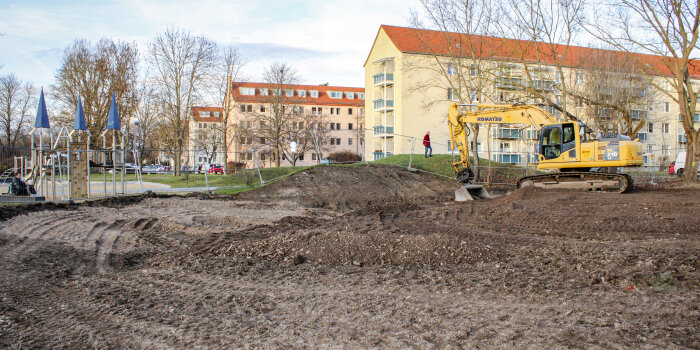 Ein Bagger steht auf einem frisch bereinigten Gelände. Rechts ist ein Spielplatz, im Hintergrund stehen Wohnblocks.