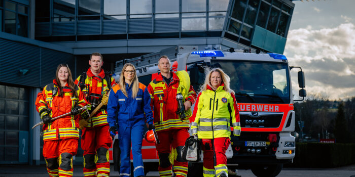 Fotomontage: Drei Frauen und zwei Männer in verschiedenen Uniformen laufen vor einem Feuerwehrfahrzeug. 