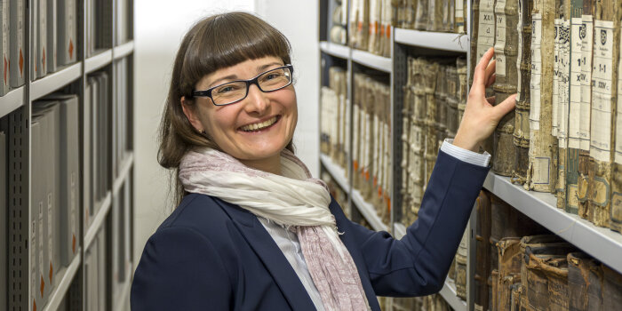 eine Frau mit Brille und braunen Haaren steht lachend an einem Regal mit alten Büchern