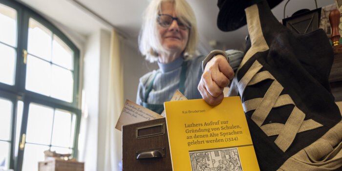 Eine Frau mit weißen Haaren ist unscharf zu sehen. Im Vordergrund stellt sie ein gelbes Buch auf. 