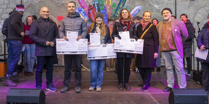 sechs Personen auf einer Bühne, drei davon halten symbolische Schecks in der Hand