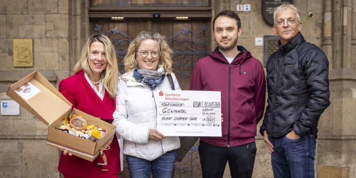 vier Personen, eine hält eine geöffnete Papp-Box, eine einen symbolischen Scheck