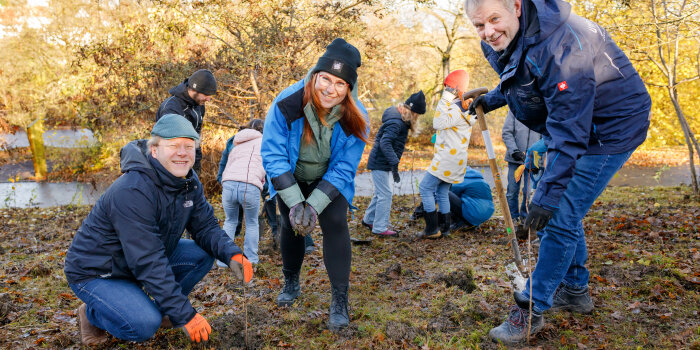 Drei Personen stehen auf einem Feld und halten einen jungen Baum und Geräte zum Einpflanzen in der Hand