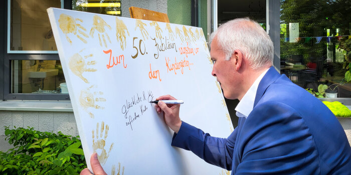 Ein Mann im Anzug schreibt auf eine Tafel Glückwünsche für einen Kindergarten. 