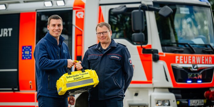 Zwei Männer posieren vor einem Feuerwehrauto mit einer leuchtend gelben Tasche. 
