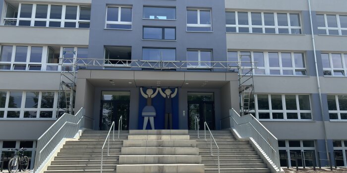 Eingang zu einer Schule. Die Fassade ist blau-grau und zwei große Figuren säumen das Portal. 