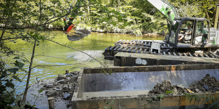 Ein Bagger steht in einem Fluss und hebt Sedimente aus, die in einen Container geschaufelt werden