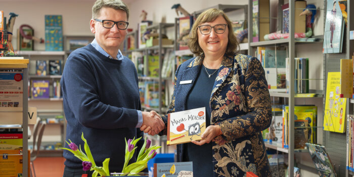 Ein Mann und eine Frau, beide tragen Brille, geben sich die Hand in einer Bibliothek. Die Frau hält ein Buch. 