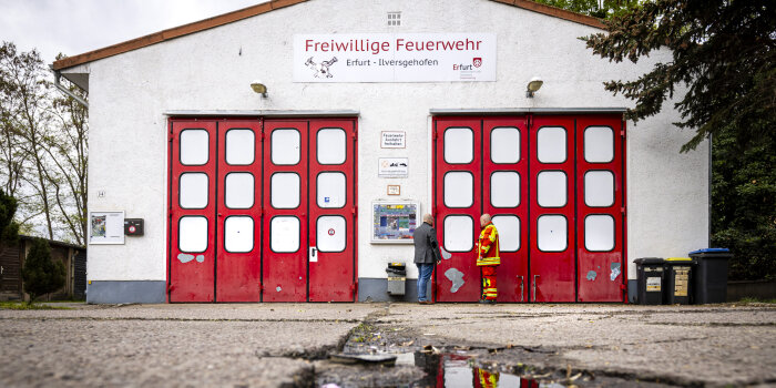 Vor einem Feuerwehrgebäude stehen zwei Männer, eine in zivil und einer in Feuerwehruniform. 