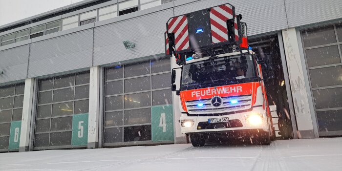 Ein Leiterwagen der Feuerwehr fährt aus einer Garage. Alle Tore sind nummeriert. Es liegt Schnee und es schneit.