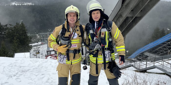 Zwei Männer stehen in Feuerwehruniform im Schnee. Im Hintergrund ist eine Skisprungschanze zu sehen. 