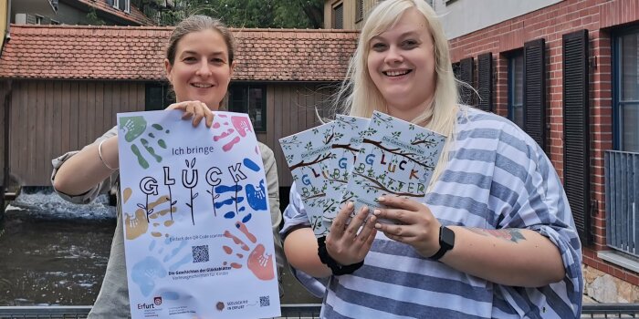 Zwei Frauen zeigen ein Plakat und mehrere Hefte mit dem Aufdruck „Glück“.