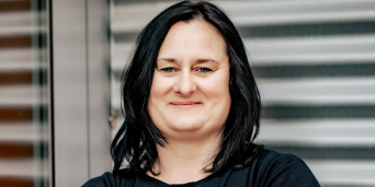 Eine Frau mit schwarzen Haaren trägt schwarze Kleidung. Links ist ihr Spiegelbild zu sehen. 