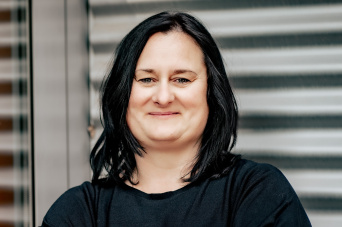 Eine Frau mit schwarzen Haaren trägt schwarze Kleidung. Links ist ihr Spiegelbild zu sehen. 