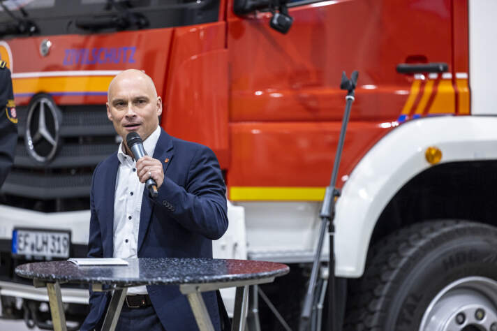 Ein Mann steht mit Mikrofon vor einem Feuerwehrfahrzeug und hält eine Rede. 