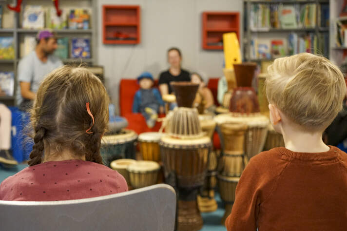 Rückansicht zweier Kinder. Sie schauen auf Trommeln, die in einer Bibliothek stehen. 