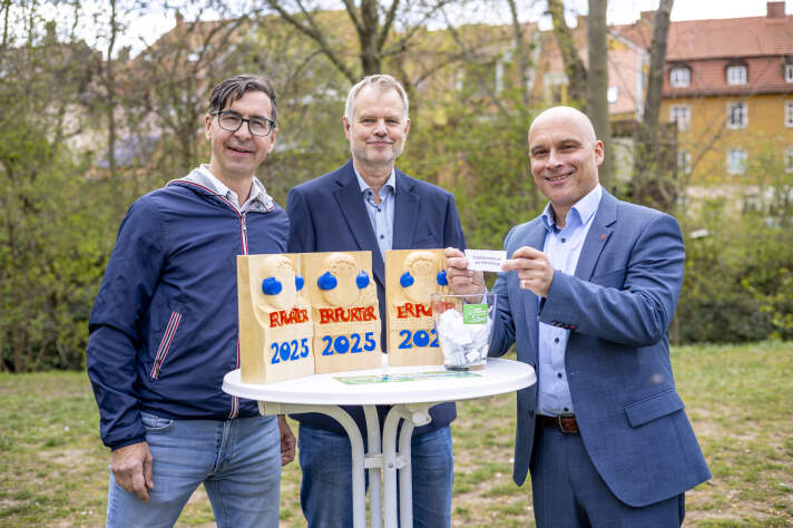 Drei Männer an einem weißen Stehtsich, einer hält einen Zettel nach oben