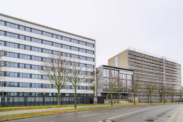 Drei mehrstöckige Gebäude. Eins davon ist eingerüstet. Davor verläuft eine Straße.