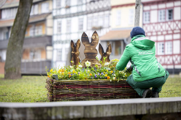 ein kleines Blumenbeet mit Osterdeko, daneben kniet eine Person in Arbeitskleidung und rückt Pflanzen zurecht