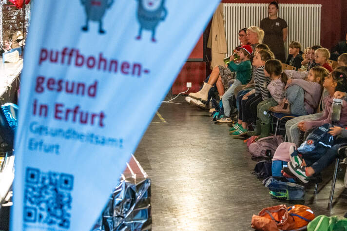 Kinder sitzen vor einer Bühne. Im Vordergrund eine Beachflag mit Aufschrift: „puffbohnengesund in Erfurt“. 