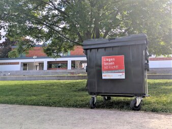 Ein großer schwarzer Müllcontainer steht an einem Parkweg vor einem großen Baum, im Hintergrund ein Gebäude