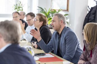ein Mann sitzt unter mehreren Menschen am Tisch, spricht und gestikuliert mit der rechten Hand