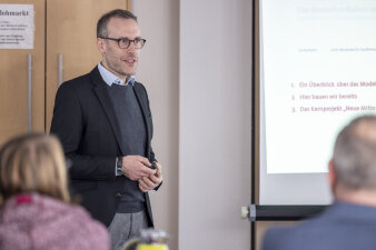 ein Mann mit Brille steht referierend neben einer Leinwand, auf der eine Präsentation läuft