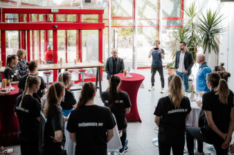 ein Mann steht im Foyer eines Gebäudes an einem Stehtisch, rundherum Menschen, einige mit T-Shirt "Thüringer HC"