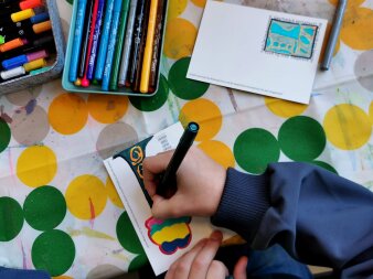 Die Hand eines Kindes hält einen Stift. Es entsteht ein Bild. Auf dem Tisch stehen Stifte und andere Kunstwerke. 