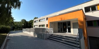 Ein zweistöckiges Gebäude mit Treppenaufgang. Das Portal ist orange gestrichen. Das Gebäude selbst ist grau. 