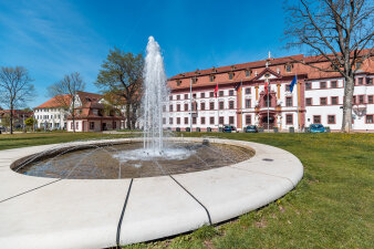 ein runder Springbrunnen vor einem langgezogenen Gebäude mit rot-weißer Fassade