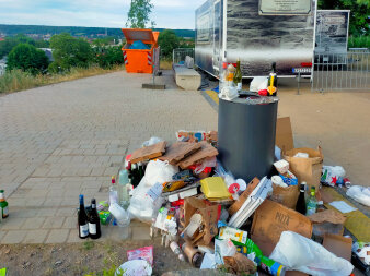 viel Müll liegt rund um einen öffentlichen Mülleimer, im Hintergrund steht ein orangefarbener Müllcontainer