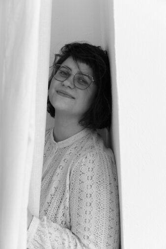 eine Frau mit kinnlangen dunklen Haaren und einer Brille steht an einem weißen Vorhang