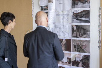 zwei Personen betrachten Fotografien und Grundrissen, die auf Papier an einer Wand hängen