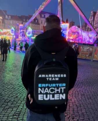 Foto: Die Erfurter Nachteulen beim Altstadtfrühling © Stadtverwaltung Erfurt Ein Mann mit beleuchtetem Rucksack und der Aufschrift Nachteulen vor bunter Rummelkulisse