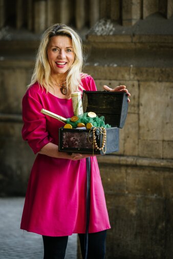 eine Frau mit blonden Haaren und einem pinken Kleid hält eine geöffnete Holztruhe