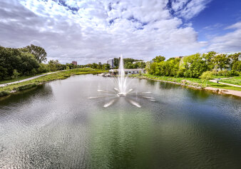 Ein Teich in einer grünen Umgeb und mit Wegen, in der Wassermitte eine Wasserfontäne
